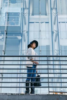 Stylish woman in a hat and fur-trimmed sweater stands by a modern glass building, exuding