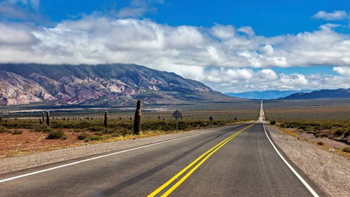 Long road leads through a desert-like landscape with mountains and cacti under a cloudy sky, The