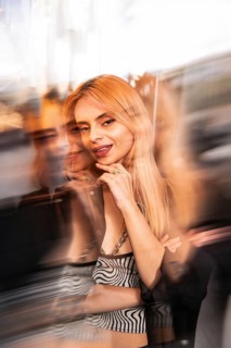 Trendy woman with long blonde hair poses confidently against a dynamic, blurred cityscape backdrop.