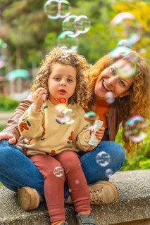 Vertical portrait of a cute blonde girl and mother sitting on a park blowing bubbles