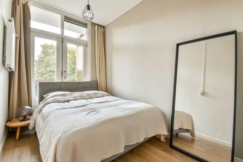 A stylish, serene bedroom featuring a bed with soft bedding, a large mirror, and bright natural