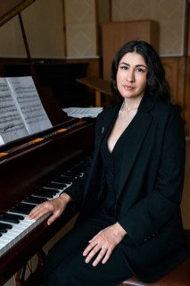 A musician in formal black attire sits beside a grand piano Sheet music is visible in the