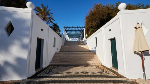 A wide staircase, flanked by white minimalist walls, leads to the top, surrounded by palm trees,