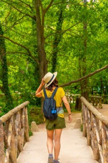 A woman wearing a straw hat and a green backpack is walking across a bridge. The bridge is
