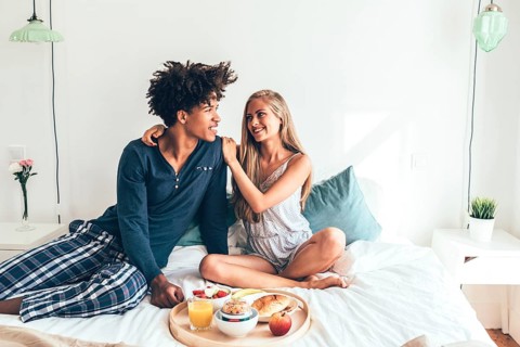A young couple sits cheerfully on a bed, sharing a breakfast tray filled with fruit, juice, and