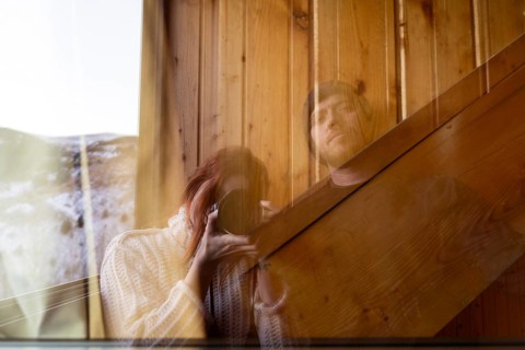 A woman with red hair holding a camera and a man are reflected in the glass window of a rustic