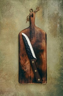 A rustic wooden cutting board displays a sharp knife resting diagonally. The textured background
