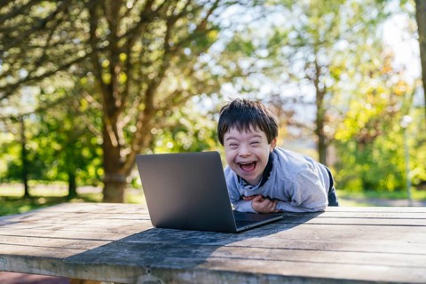 A cheerful child with Down syndrome is smiling joyfully at a laptop while sitting at a wooden table