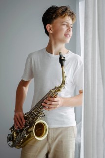A young saxophonist in a white shirt looks out the window, standing in soft sunlight. The relaxed