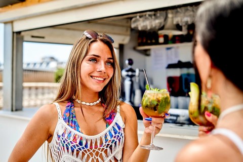 Two friends are at a vibrant beach bar during a sunny summer day, holding colorful cocktails and