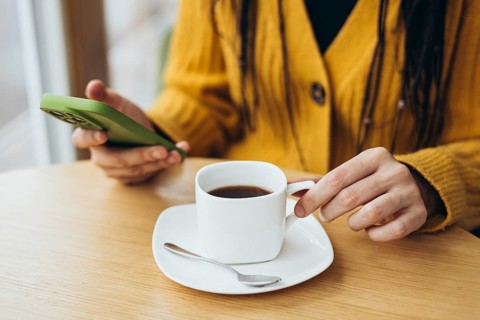 A person in a yellow sweater holds a smartphone while enjoying coffee at a wooden table A white