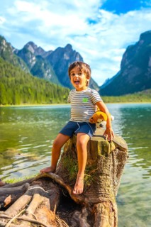 Carefree child sticking out tongue, sitting on tree stump by lake dobbiaco with plush toy, enjoying