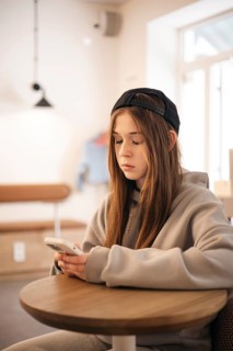 A teenage girl wearing a hoodie and cap sits at a small table, engrossed in her smartphone. The