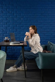 A woman in casual attire sips tea, seated at a table with a laptop, against a vibrant blue brick