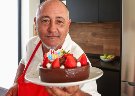 A cheerful senior man in a red apron holds a chocolate cake adorned with strawberries and a Happy
