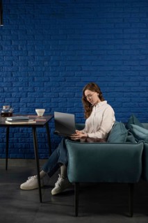 A woman in casual attire works on a laptop, seated on a teal sofa against a blue brick wall. A