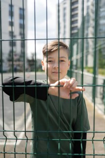 A young boy in a dark shirt stands pensively behind a green wire fence, with urban high-rise