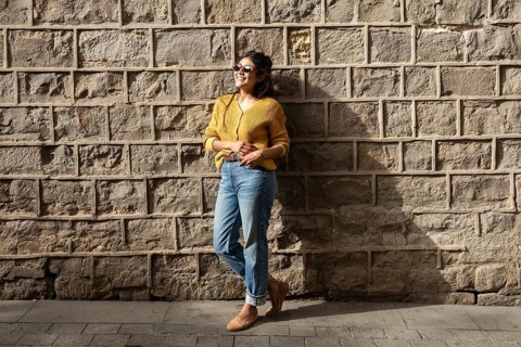 A Latin woman in casual attire, wearing sunglasses, smiles while leaning against a sunlit brick