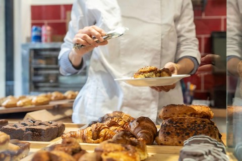 Chef wearing a white uniform carefully serving a fresh cinnamon roll onto a plate with tongs.