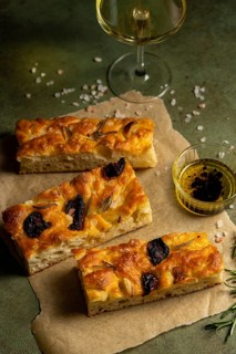 Freshly baked homemade focaccia topped with garlic and olives served on parchment paper.