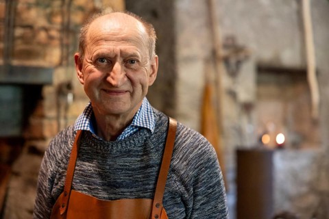 A skilled blacksmith wearing a leather apron smiles warmly in his rustic workshop. The background