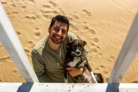 A cheerful man holding his dog on a sunny beach in Greece. The sand is marked with footprints.
