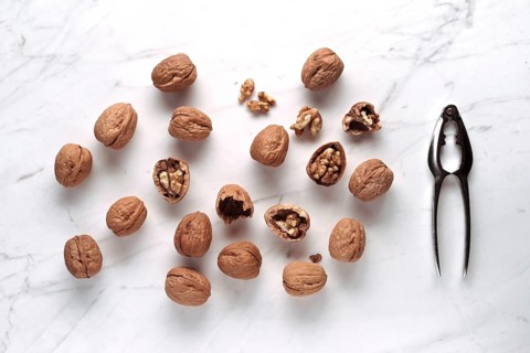 Whole and cracked walnuts scattered on a marble surface, next to a shiny metal nutcracker. The