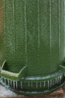 Upside down green PVC and rubber thrash can covered with thick sheet of ice and hanging icicles