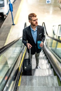 Businessman going up escalator in subway station, holding reusable coffee cup and pulling trolley