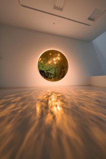 A round mirror on the wall casts warm light reflections onto the floor in an art space, Schauwerk