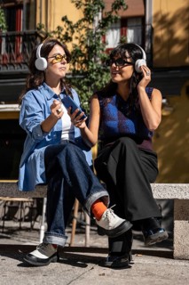 A multiethnic lesbian couple sits on a bench outdoors, wearing headphones and sharing a joyful