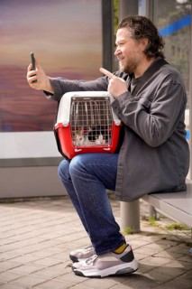 A man sits at a bus stop with two cats in a carrier on his lap, taking a selfie with his smartphone