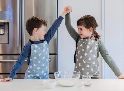 Two young siblings in star patterned aprons enthusiastically raise their hands in celebration while