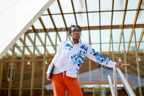A man in a vibrant, floral shirt and orange pants poses confidently near a modern glass building,