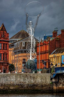 Modern metal sculpture on a riverbank in front of historic buildings under a cloudy sky, Belfast