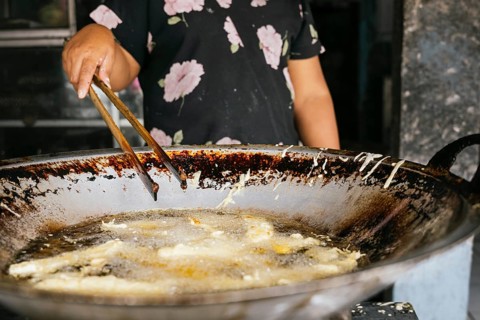 A woman prepares street food in a large wok using tongs. The image captures the sizzling oil and