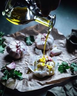 Close-up of garlic bulbs drizzled with olive oil, surrounded by fresh herbs on crumpled parchment