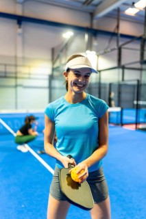 Young woman smiling and holding a pickleball paddle with a perforated ball, standing on a bright