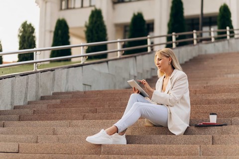 A businesswoman in casual attire sits on outdoor steps with a digital tablet, concentrating on her