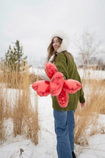 A woman wearing a green sweater and fur hat holds red heart shaped balloons in a snowy park. She
