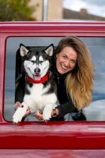 A joyful woman with long hair leans out of a red car window, holding a happy husky dog wearing a
