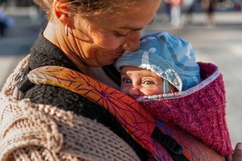 A tender moment between a mother and her baby in a colorful carrier. The warm sunlight illuminates