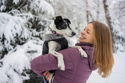 A woman in a purple jacket holds her dog lovingly in a snowy forest. Her smile and the snowy scene