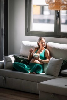 Pregnant woman in a green yoga outfit relaxes on a sofa, enjoying a healthy bowl of fruit. Sunlight