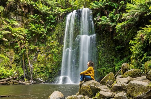 A woman sits peacefully on moss covered rocks near a waterfall surrounded by lush green ferns in