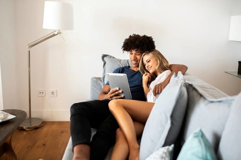 A young couple relaxes on a cozy couch, sharing a tablet. The scene captures a moment of joy and