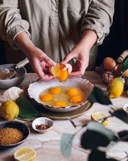 A person cracks eggs into a bowl on a table surrounded by lemons, brown sugar, and spices,