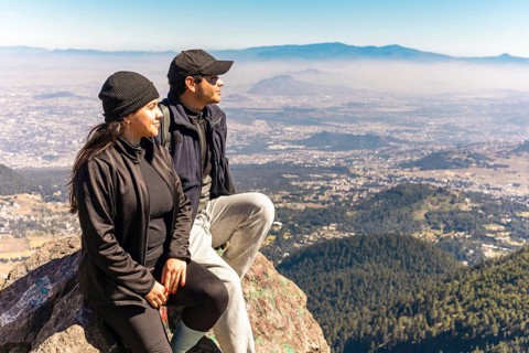 Two friends are seated on a mountain rock, gazing at a stunning panoramic view They are bundled up