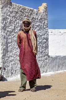 A man in traditional Tuareg clothing stands in front of a sunlit, plastered wall, Local people in