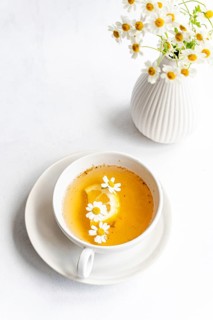 Top view of a white cup containing tea with daisies and lemon slice on a saucer. Nearby, a white